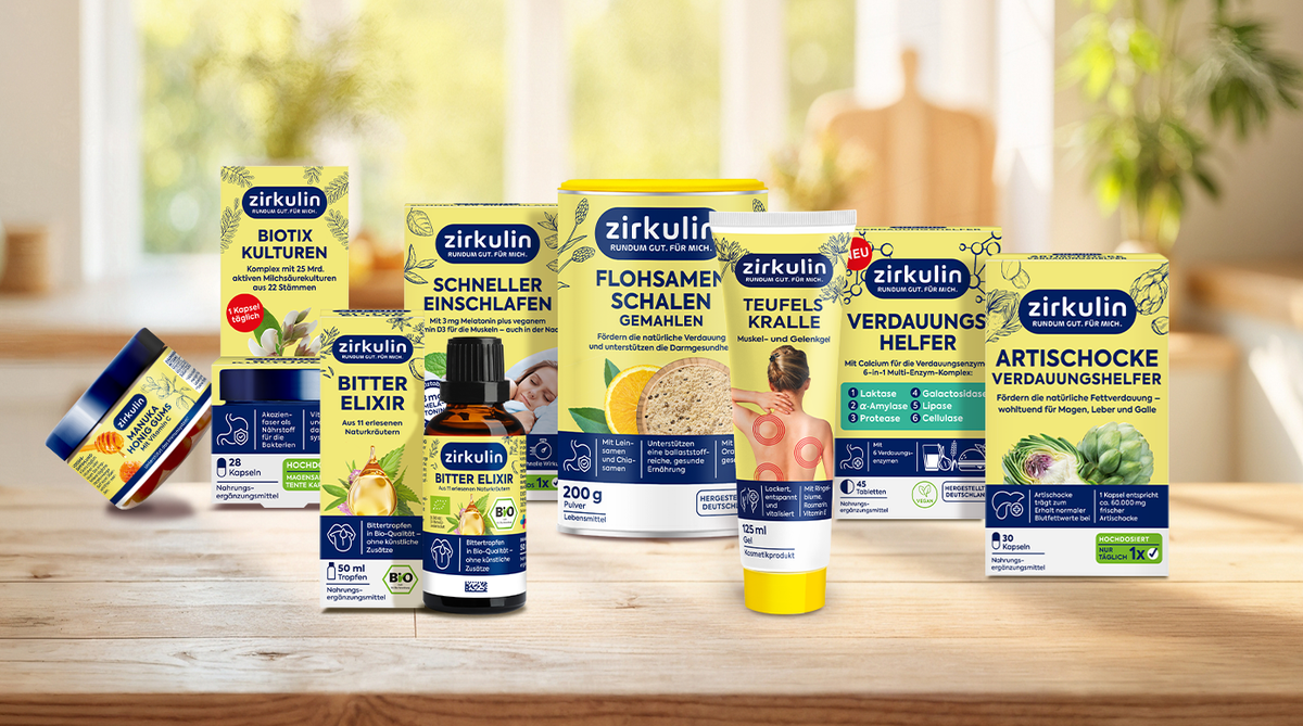 Verschiedene Zirkulin-Gesundheitsprodukte, darunter Nahrungsergänzungsmittel, Cremes und Pulver, sind auf einem Holztisch vor einem unscharfen Innenraumhintergrund ausgestellt. Die Verpackungen sind überwiegend in Gelb und Blau gehalten und mit Pflanzenmotiven verziert.